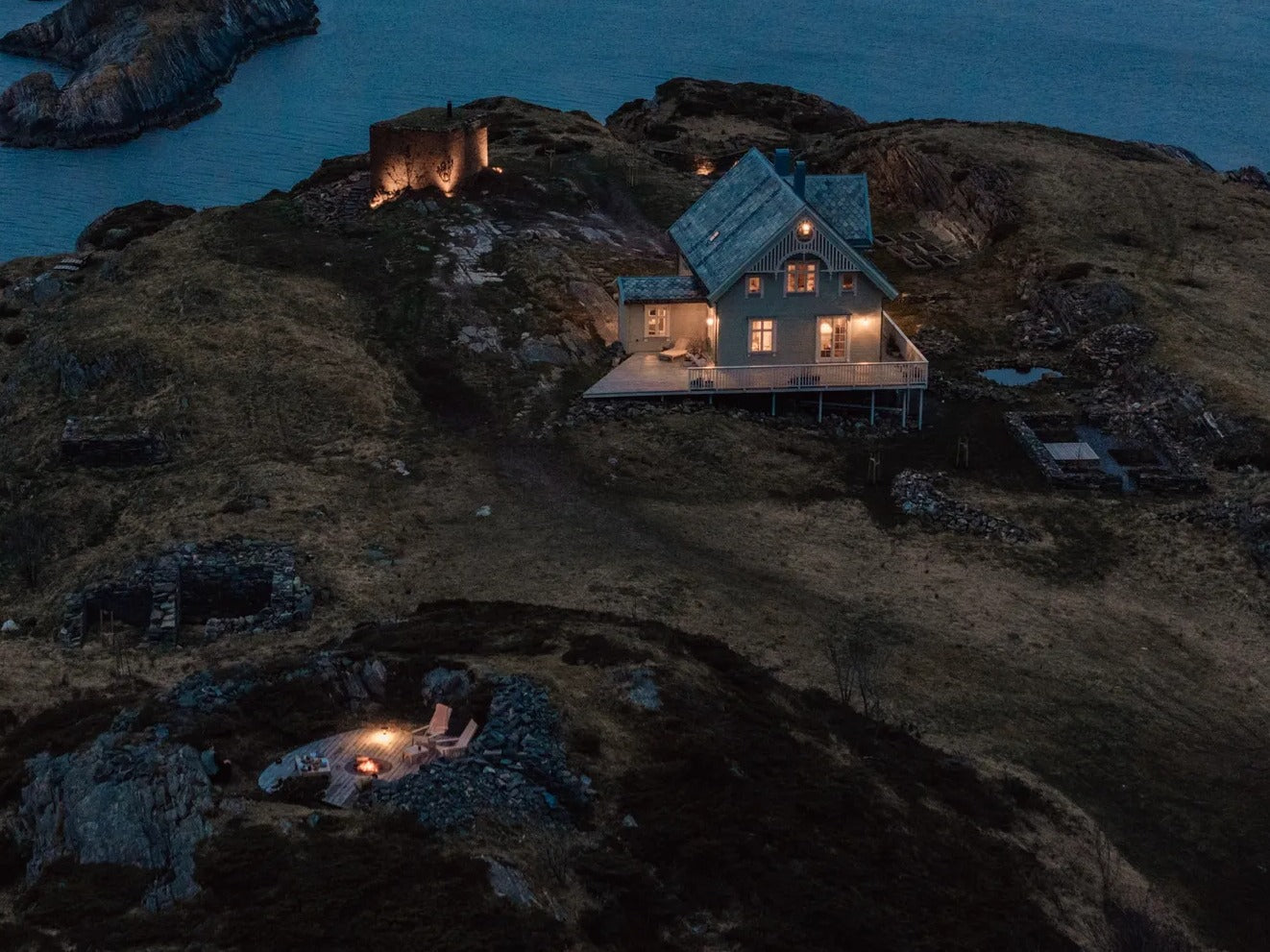 Cozy house on rocky coastline at dusk with outdoor fire pit, relaxing chairs, and sea view