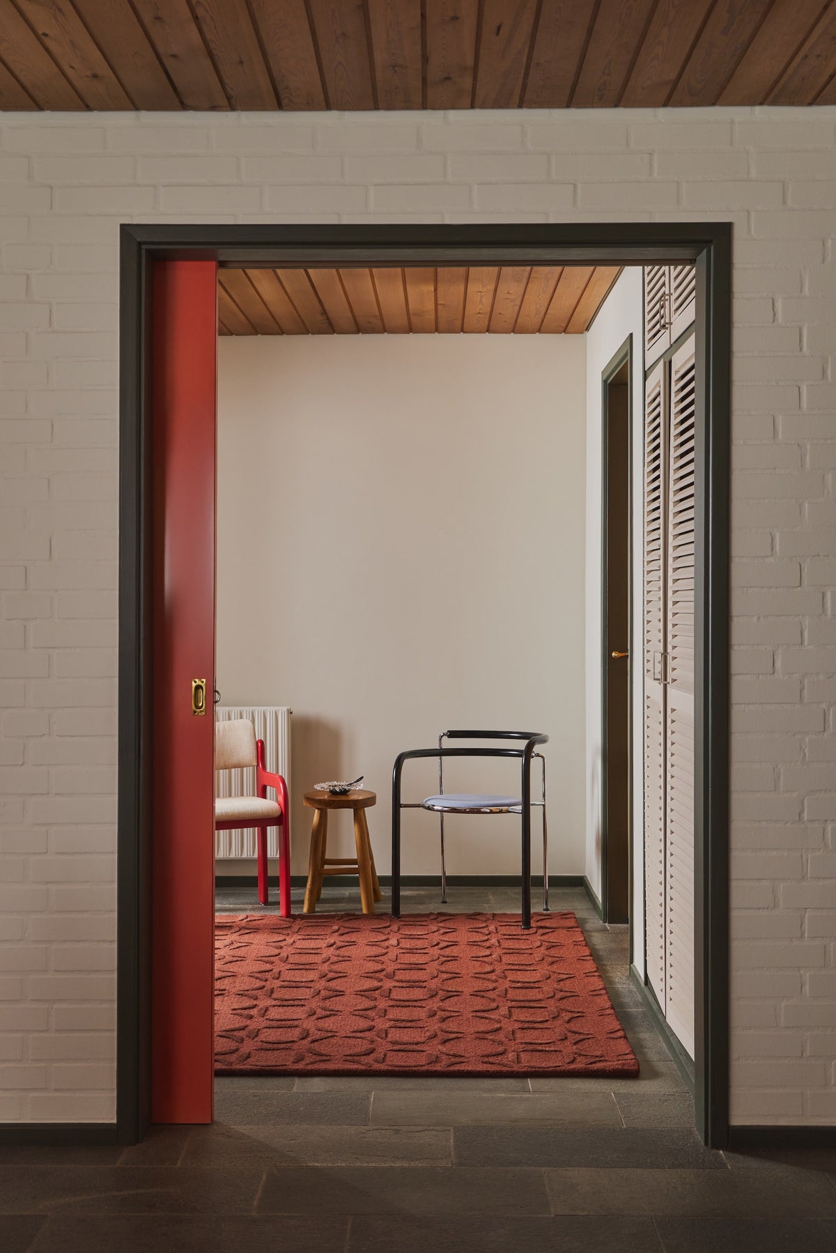 A view through a dark-framed doorway into a hallway or room with a red patterned rug, a red chair, a small wooden stool, and a black metal chair, set against a white brick wall and wood ceiling.