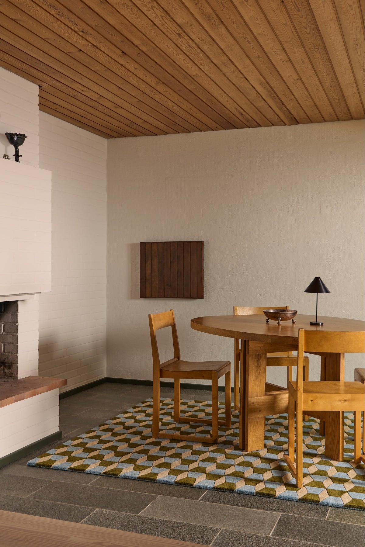 A mid-century modern dining room featuring a round wooden table and four matching wooden chairs on a geometric patterned rug. The room has textured off-white walls, a wood-paneled ceiling, and a stone fireplace on the left.
