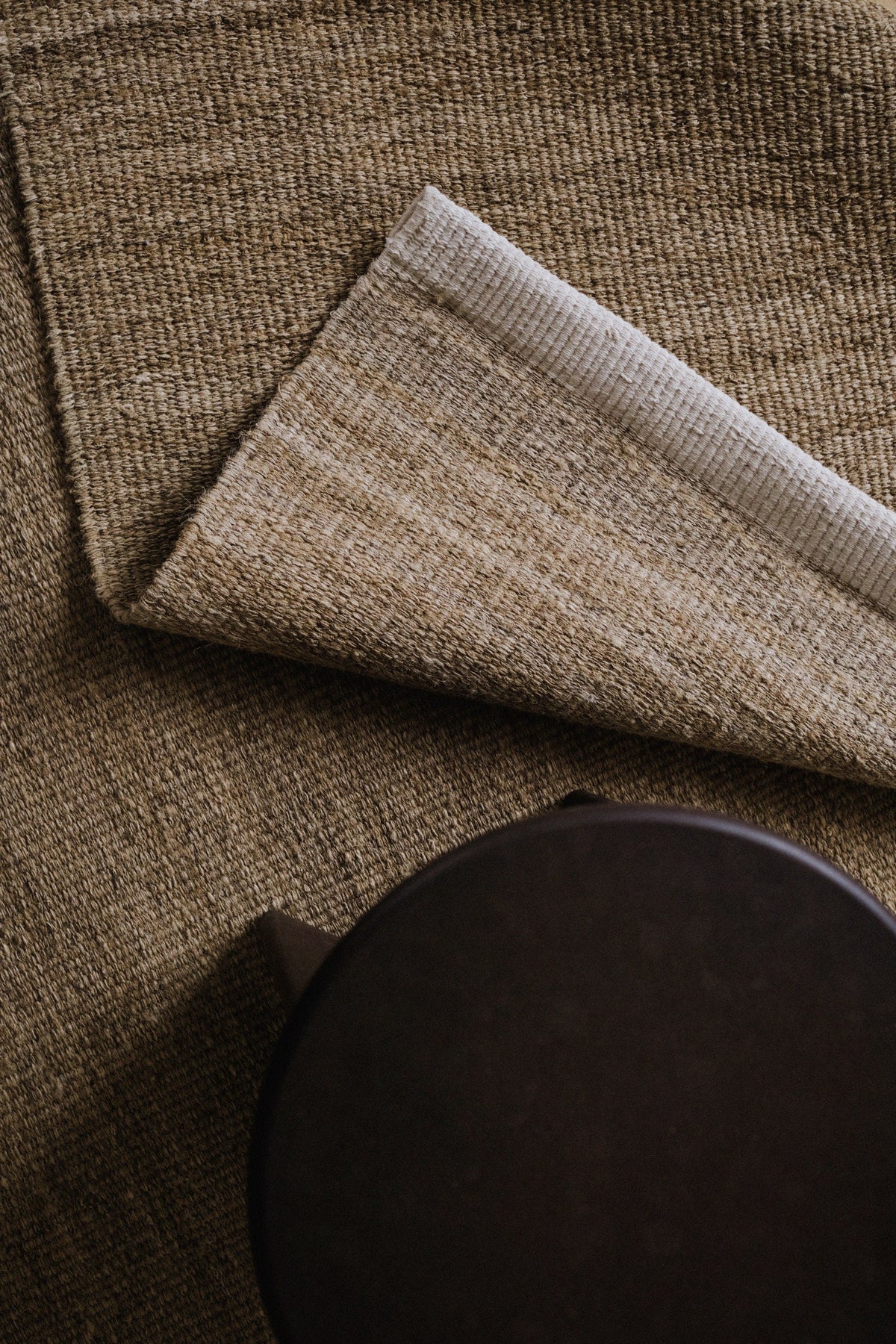 A close-up of a textured woven rug in warm taupe and beige tones, with one corner folded back to reveal the pile and backing, next to a dark round stool.