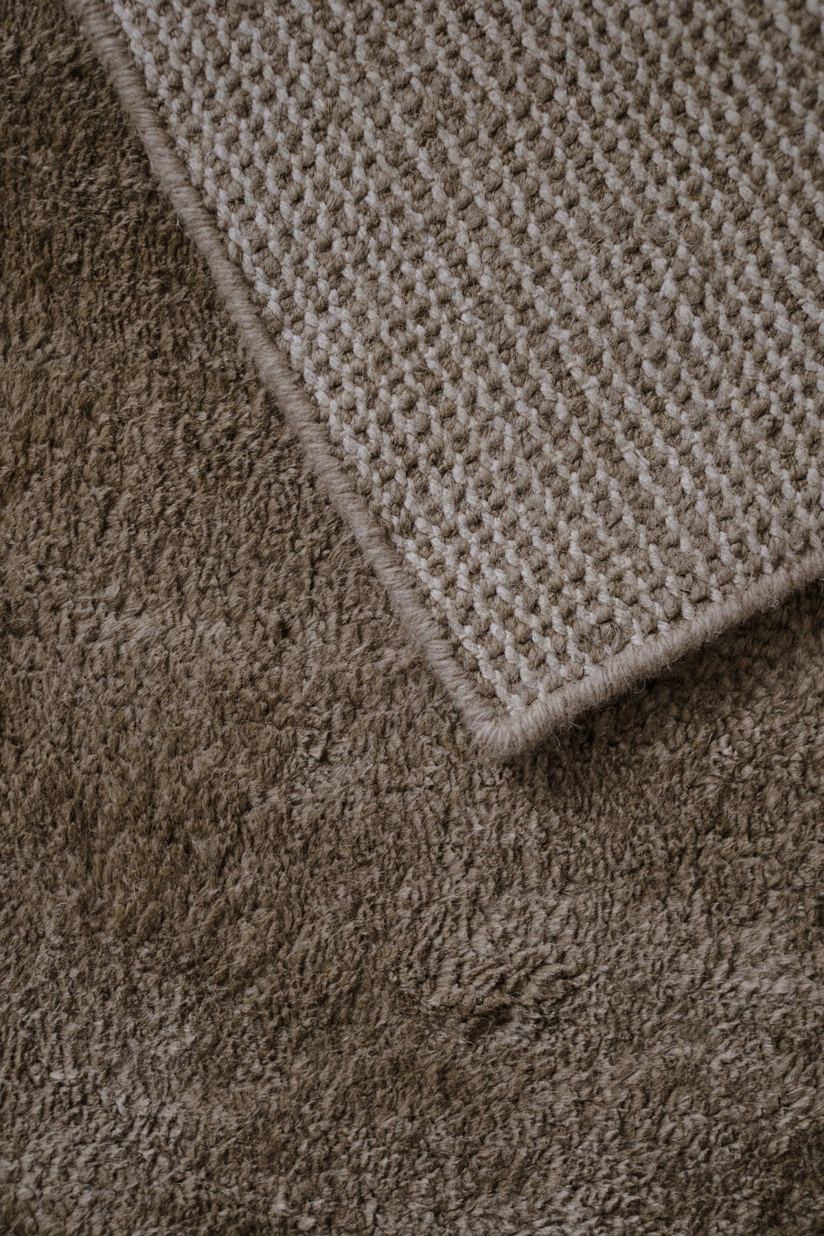 Close-up of a plush taupe-brown rug with a contrasting light beige woven texture and stitched edge.